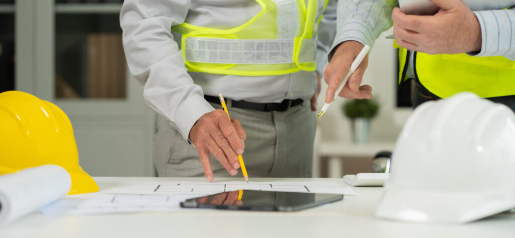 Contractors pointing at blueprints on table with tablet and construction hats surrounding it